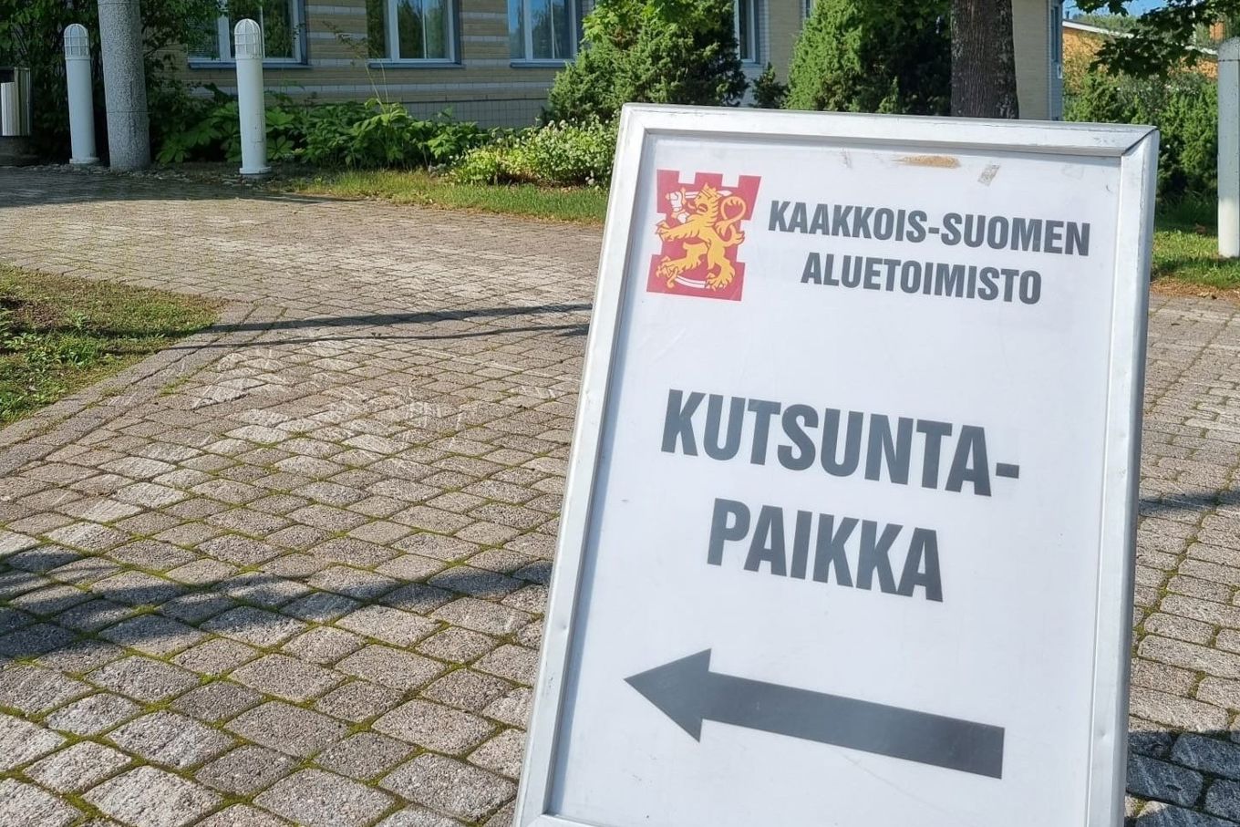 Taipalsaaren kunnantalon edustalla iso kyltti, jossa teksti Kaakkois-Suomen aluetoimisto Kutsuntapaikka. Tekstin alla on musta nuoli, joka osoittaa kunnantalon sisäänkäynnille.