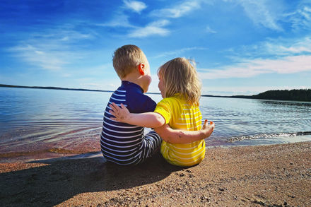 Kaksi lasta istuu hiekkarannalla ja pitää toisistaan kiinni.