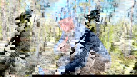 Mies kyykyssä metsäkalliolla. Mies näyttää kameralle peukkua.