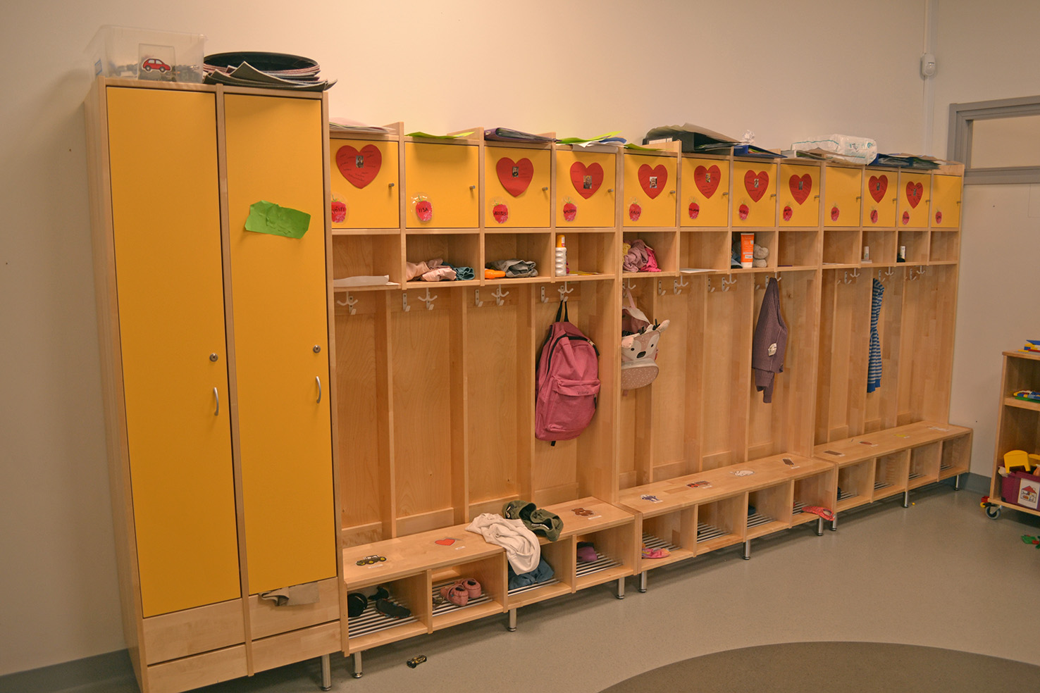 Keltainen naulakko päiväkodin aulassa. Naulakossa roikkuu lasten vaatteita ja reppuja.