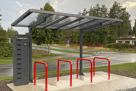 Harmaa pyöräkatos, jonka alla on neljä punaista pyöräkaarta. Kaarien vieressä korkea harmaa latauskaappi.